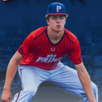 The image features a baseball player wearing a red USA PRIME jersey with white lettering and blue accents, along with a blue cap bearing a red "P" logo. He is in a ready defensive stance, likely as an infielder, with a focused expression. The background has a blue overlay with a faint cityscape or stadium visible.