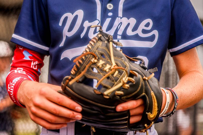 This image features a baseball player wearing a USA Prime jersey, holding a black and gold baseball glove. The detailed close-up highlights the stitching and lacing of the glove, as well as the player's red compression sleeve and wristband. The USA Prime branding on the uniform suggests an affiliation with the USA Prime Baseball organization, which is known for its high-level travel baseball teams and player development programs.