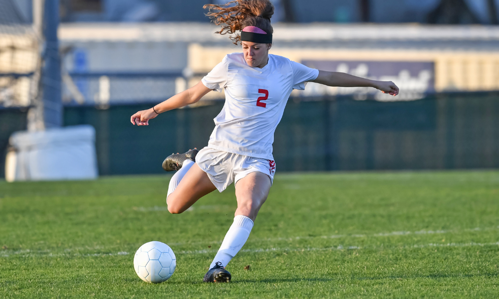 A female soccer player wearing a white jersey with the number 2 is captured mid-kick, striking a white soccer ball on a green grass field. Her hair flows behind her as she follows through on the kick, with a focused expression and an athletic stance.