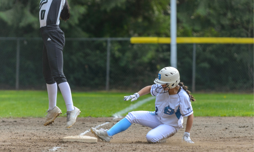 A softball player in a white and light blue uniform slides into a base, kicking up dirt as she reaches for safety. An opposing player in a black and white uniform leaps into the air, attempting to catch the ball for a tag. The background features a fenced field with green grass and trees.