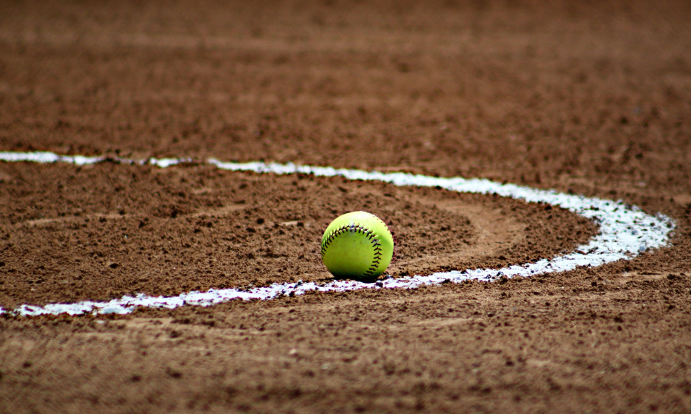 A bright yellow softball rests on the dirt infield near a freshly chalked white foul line. The textured surface of the field and the curve of the line add depth to the image, capturing the essence of the game.