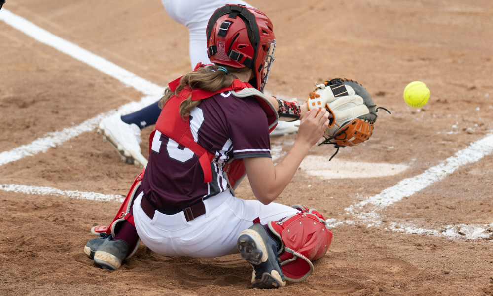 A softball catcher in maroon and white protective gear crouches behind home plate, reaching out with her glove to catch a fast-approaching yellow softball. A baserunner in white cleats slides past the plate, adding to the action on the dirt field.