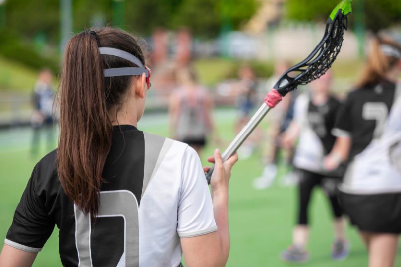 This image captures a female lacrosse player from behind, wearing a black and white jersey with the number 0. She holds her lacrosse stick over her shoulder while standing on a green artificial turf field. Other players, also dressed in similar uniforms, are visible in the background, engaged in practice or warm-up activities. The setting suggests a team practice session or a pre-game preparation, emphasizing the player's focus and readiness for competition. The headband and ponytail add to the athletic look, highlighting the intensity and energy of the sport.