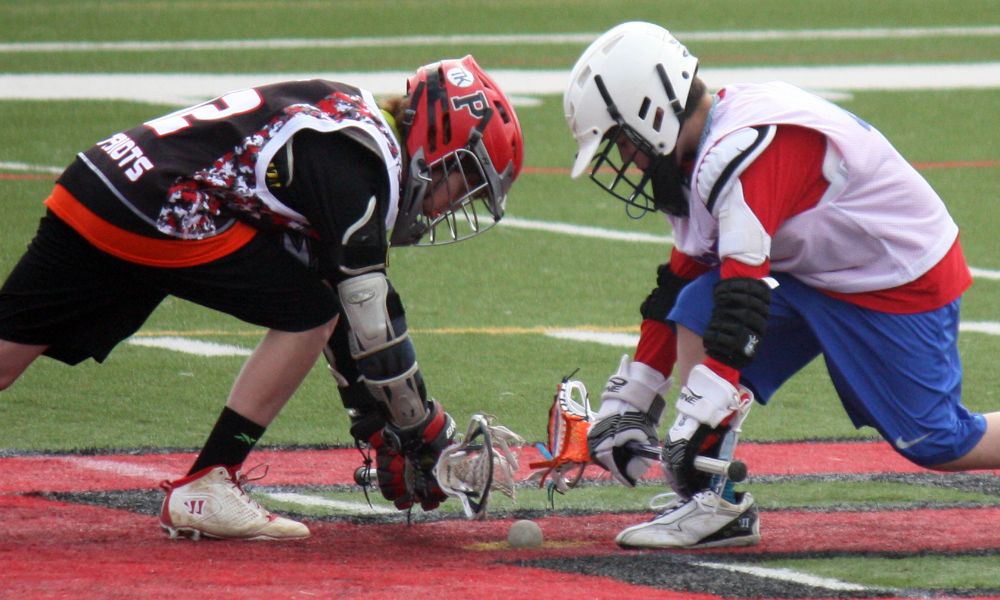 This image captures an intense faceoff in a lacrosse match, featuring two young athletes in full gear. The player on the left is wearing a red and black uniform with a matching red helmet, representing a team with "Patriots" on the jersey. The player on the right is in a white and red uniform with a white helmet. Both are crouched low, sticks pressed against the turf, battling for possession of the ball. Their protective gloves, elbow pads, and helmets indicate a high level of competition and safety awareness. The synthetic turf field with bold red and black markings adds to the competitive atmosphere of the match.