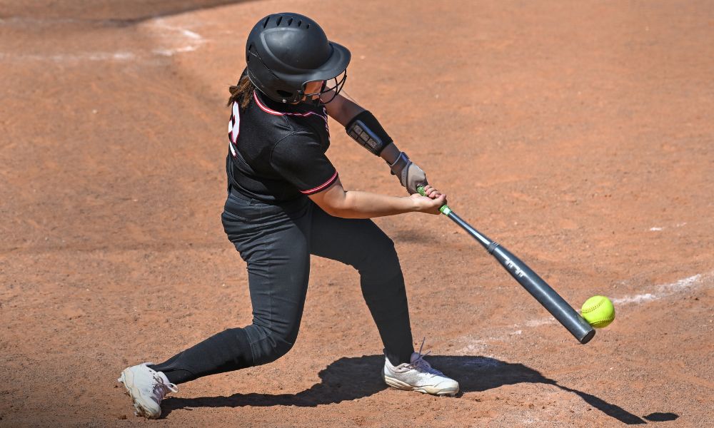 This image captures a softball player in action, wearing a black uniform with red accents. She is making contact with the bright yellow softball while gripping a dark-colored bat. Her stance is strong, with bent knees and a forward-leaning posture, demonstrating focus and power. The black helmet with a protective faceguard ensures safety as she swings. The dusty infield and clear lighting suggest a well-lit, outdoor game environment. This moment encapsulates the intensity and precision required in competitive softball.