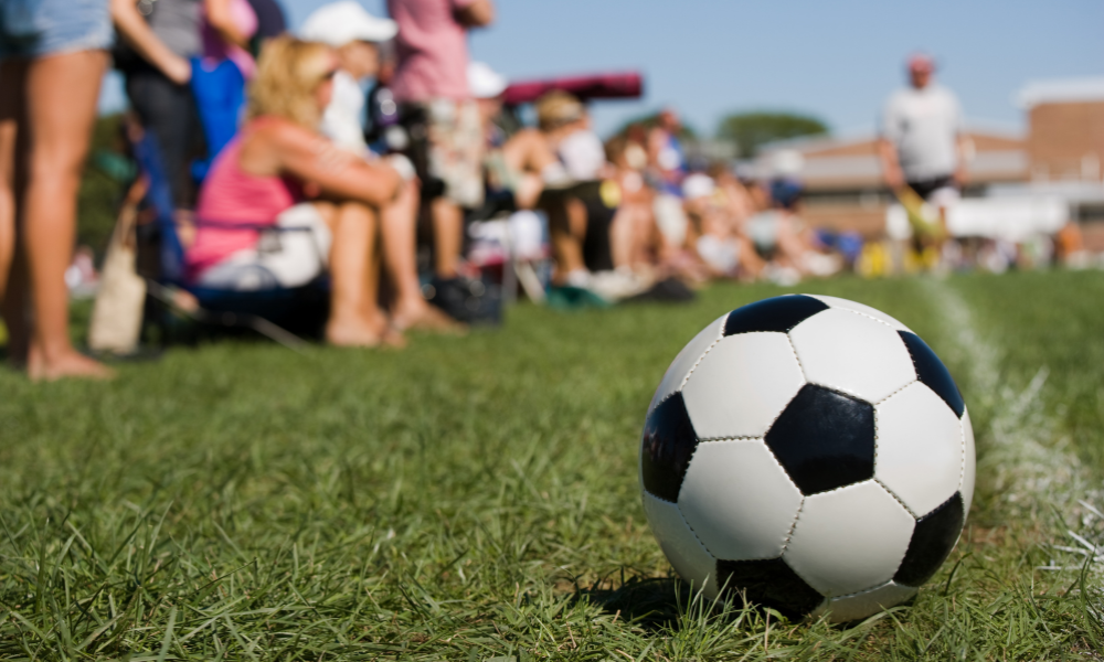 A black-and-white soccer ball sits on the green grass near the sideline of a youth soccer field. In the background, spectators sit on folding chairs, watching the game under a bright blue sky.