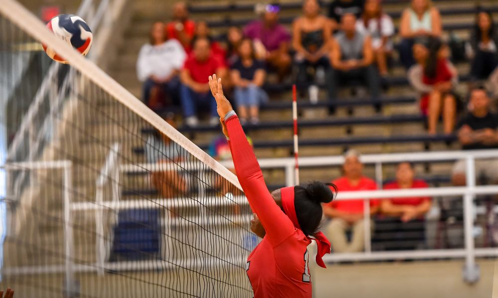 The image showcases a volleyball player in a red uniform leaping high to make contact with the ball at the net. Her outstretched arm and focused posture suggest an intense moment in the game, likely a block or an attempt to return the ball. The background features a crowd of spectators seated in the stands, attentively watching the match. The gymnasium setting, with its bright lighting and structured bleachers, adds to the competitive atmosphere.