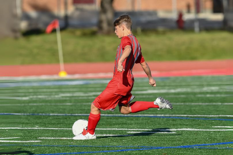 A young male soccer player wearing a red uniform is captured mid-kick on a synthetic turf field, striking a white soccer ball with his right foot. His posture is focused, with his leg extended and arms slightly spread for balance, while a red corner flag and a running track are visible in the background.