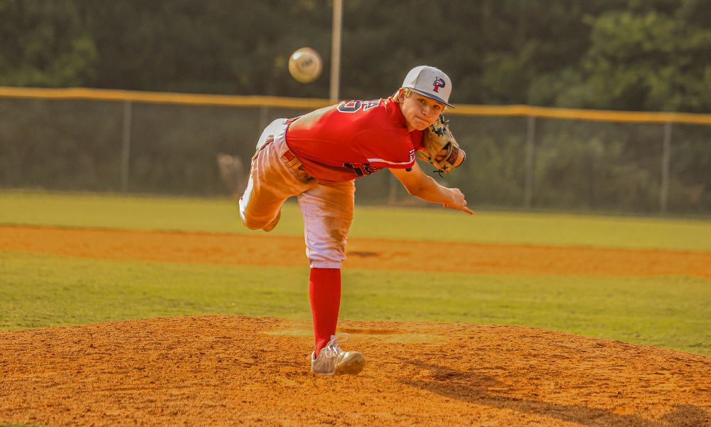 This image features a USA Prime baseball pitcher in action, wearing a red jersey and white pants with red socks. The player is captured mid-pitch, showcasing a strong, athletic stance with one leg extended and the throwing arm fully extended forward. The focus and intensity on the pitcher's face indicate concentration and precision in his delivery. The baseball is seen in motion, slightly blurred, highlighting the speed and power of the pitch. The background consists of a well-maintained baseball field, with the pitching mound and outfield fence visible, adding depth to the action. The warm lighting suggests this game is being played during the golden hour, either at sunset or early evening, which adds a dramatic effect to the scene. The composition and angle of the shot effectively capture the athleticism and energy of competitive baseball.