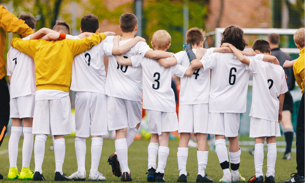 A youth soccer team in white uniforms huddles together with arms around each other's shoulders, showing unity and teamwork before a match. The players face a soccer goal on a green field, with trees and buildings in the background.