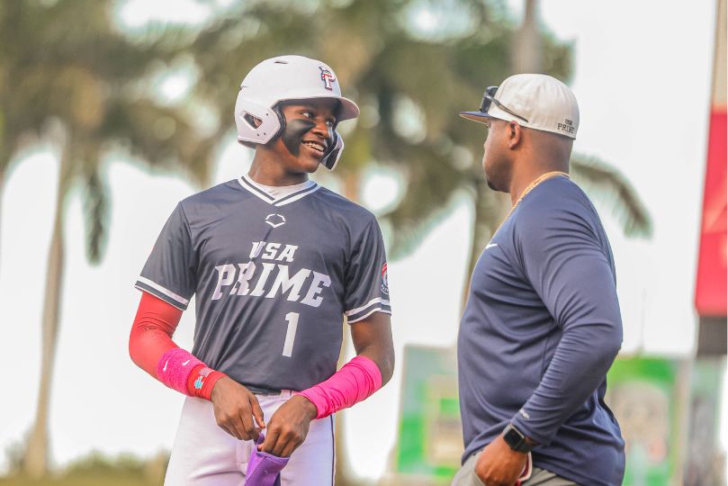 This image captures a USA Prime baseball player wearing a navy blue jersey with the number 1 and a white batting helmet, engaged in a lighthearted conversation with a coach or mentor. The player's bright pink arm sleeves and wristbands stand out, adding a touch of personality and flair to his uniform. The coach, dressed in a navy long-sleeve shirt and a white cap, appears to be offering words of encouragement or strategy. The body language of both individuals suggests a positive and engaging interaction, likely during a break in play. The background, featuring palm trees, indicates a warm-weather baseball setting, possibly in Florida or another southern state. The relaxed yet focused atmosphere in the image highlights the camaraderie and mentorship that is central to team sports like baseball.