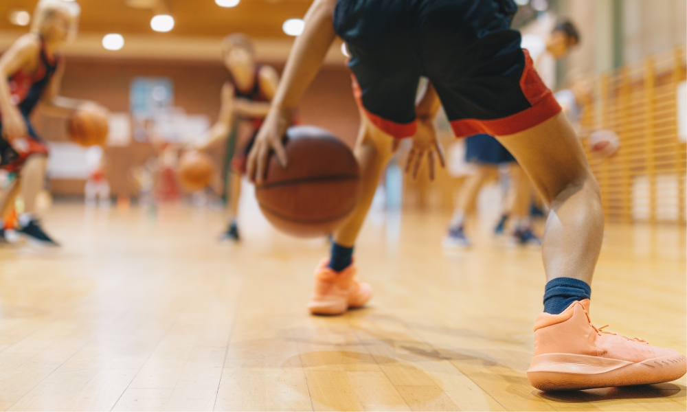 A close-up shot of young basketball players practicing dribbling drills in a gymnasium. The focus is on the legs and hands of a player wearing bright orange sneakers, controlling the ball on a polished wooden floor, while other players in the background also engage in the drill.