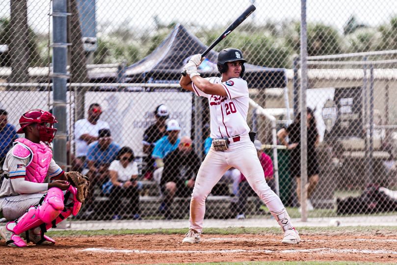 This image captures a USA Prime baseball player at bat, poised and ready to swing. The player is in a wide, balanced stance, gripping the bat with both hands, eyes locked in on the incoming pitch. His crisp white uniform with red accents and the number 20 stands out against the chain-link fence and spectators in the background. The opposing catcher, fully geared in bright pink protective gear, is crouched and focused, prepared to receive the pitch. The setting suggests a competitive game atmosphere, with engaged fans and team personnel watching from behind the fence. The overall tone of the image reflects intensity and anticipation in the game.