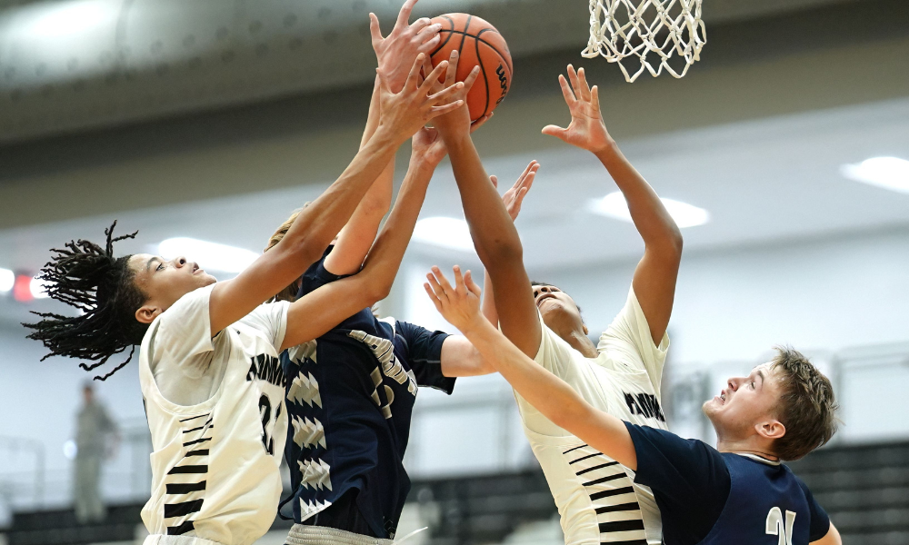 Basketball players from opposing teams in white and navy blue jerseys leap into the air, reaching for the ball in an intense rebound battle near the hoop. Their arms extend as they compete for possession, with the net and gymnasium lights visible in the background.