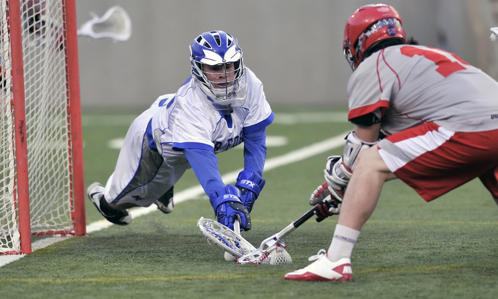 A lacrosse goalie in a white and blue uniform dives forward, fully extended, attempting to block a shot near the goal. An opposing player in a red and gray uniform, holding a lacrosse stick, is poised to strike. The intense action unfolds on a green turf field.