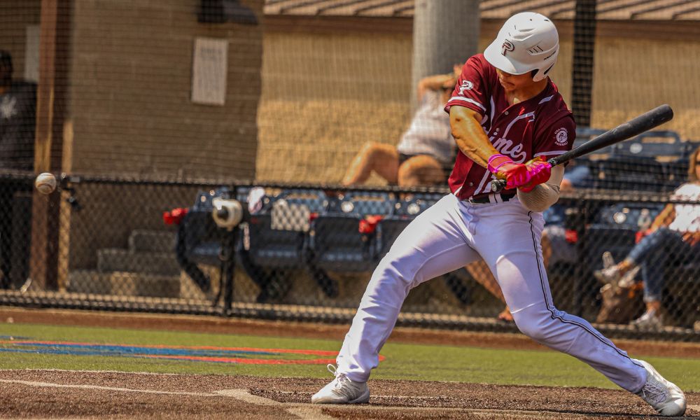 This image captures a baseball player in the midst of a powerful swing, wearing a USA Prime uniform. The batter is intensely focused on the incoming pitch, demonstrating excellent form with a strong stance and a full-body rotation. The pink batting gloves add a pop of color, contrasting with the maroon and white uniform. The background shows a fenced-off spectator area with onlookers watching the game, further emphasizing the competitive atmosphere. The lighting and framing highlight the action, making this a dynamic shot of a key moment in play.