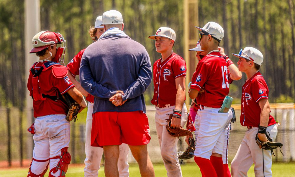 Baseball coach in a navy hoodie and red shorts giving a pep talk to a group of USA Prime players in red uniforms.