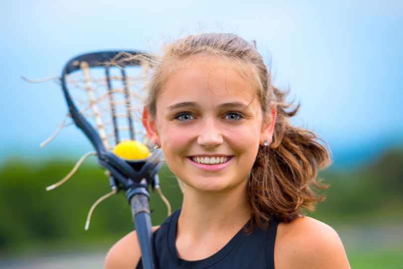 A young female lacrosse player in a black jersey smiles confidently while holding a lacrosse stick with a yellow ball in the net. The background features a blurred outdoor field with greenery and a bright sky.