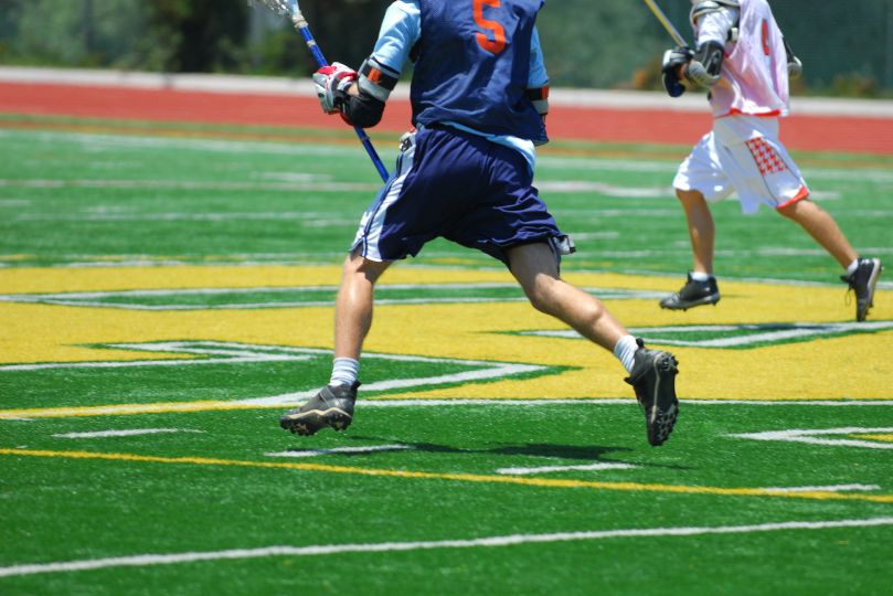 Two lacrosse players sprint across an artificial turf field, one in a navy blue jersey with the number 5 and the other in a white and orange uniform. Both players hold lacrosse sticks as they compete for possession, with their cleats digging into the vibrant green field marked with yellow lettering.