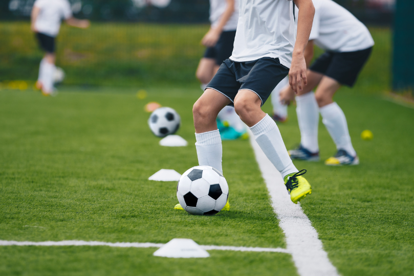 Young soccer players in white jerseys and black shorts practice dribbling drills on a green artificial turf field. They maneuver black-and-white soccer balls around small training cones, focusing on footwork and ball control.