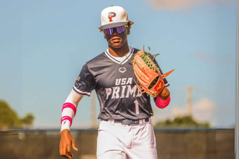 A baseball player wearing a navy blue USA Prime jersey with the number 1 walks confidently across the field. He sports a white cap with a red "P" logo, reflective sunglasses, and pink arm sleeves while holding a baseball glove in his left hand. The background features a clear sky and fencing.