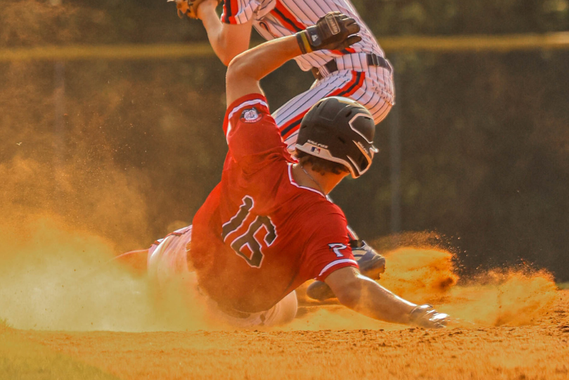 A baseball player in a red jersey with the number 16 slides into a base, kicking up a cloud of dust. An opposing player in a white pinstriped uniform attempts to make the tag, with intense action captured on the infield dirt.