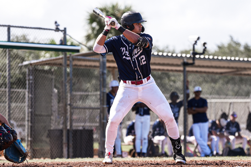 A baseball player in a navy blue jersey with the number 12 stands in the batter's box, gripping his bat and preparing to swing. He wears a black helmet, white pants, and black cleats. The catcher and umpire are partially visible, while teammates watch from the dugout in the background.