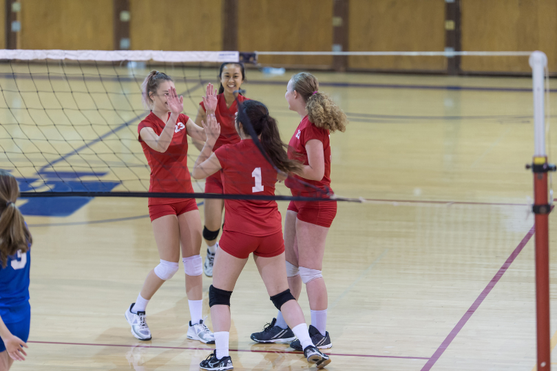 Team of female volleyball players celebrating a point in a gymnasium, wearing red uniforms and gathered near the net with high-fives and smiles.