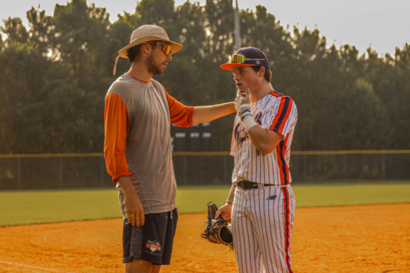 A baseball coach wearing a sun hat and sunglasses places a hand on the shoulder of a player in a white pinstriped uniform with orange and blue accents. The player, holding a glove, listens attentively on a dusty infield with trees and a fence in the background.