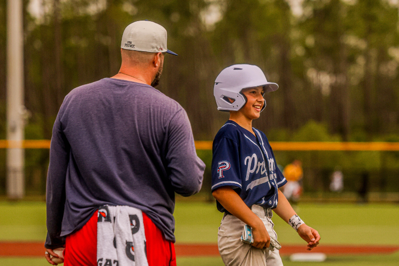 A young baseball player in a navy blue uniform and white helmet smiles while standing on the field next to a coach wearing a gray cap and blue long-sleeve shirt. The background features a baseball field with trees and a yellow fence under an overcast sky.