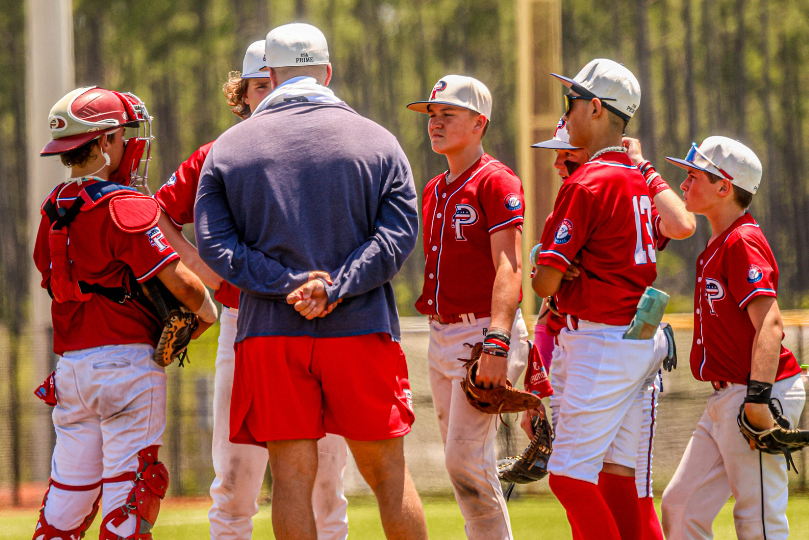 A youth baseball team in red and white uniforms huddles around their coach on the field during a game. The players, including a catcher in protective gear, listen attentively as the coach, wearing a blue sweatshirt and red shorts, gives instructions under the bright sun.