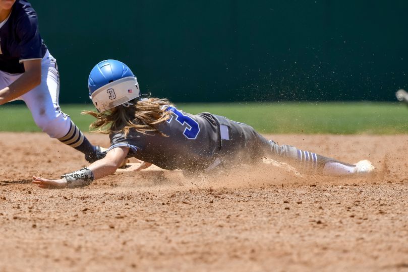 A softball player in a blue uniform and helmet slides headfirst into a base, kicking up a cloud of dust. An opposing player in a navy blue and white uniform reaches down in an attempt to tag her out. The action takes place on a sunlit dirt infield with a green outfield fence in the background.