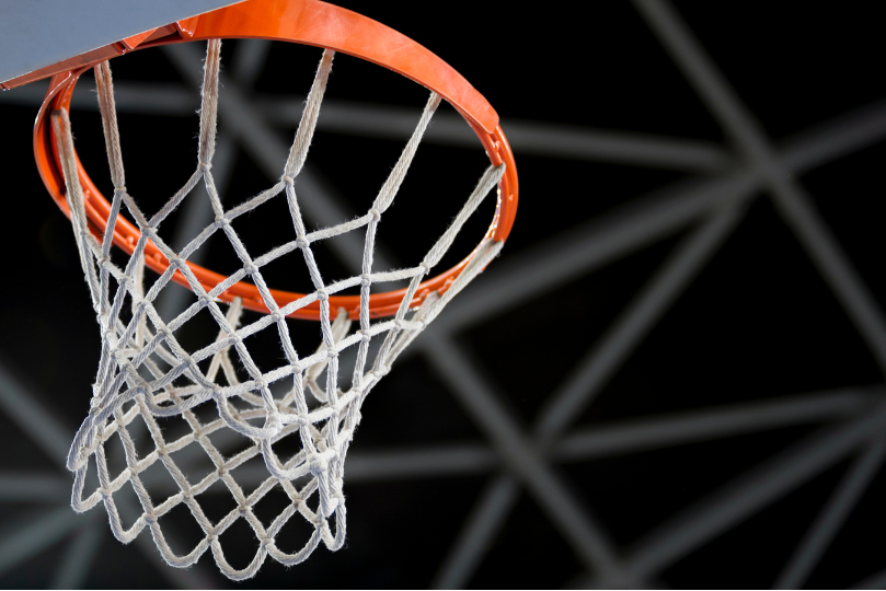 A close-up of a basketball hoop with a white net hanging from an orange rim, set against the dark ceiling structure of an indoor arena. The metal framework in the background adds depth to the image.