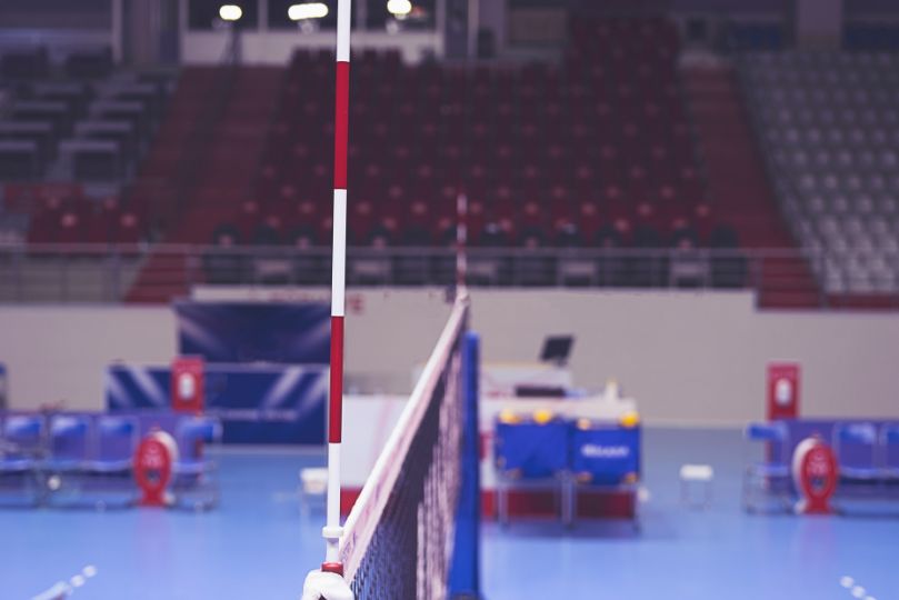 A close-up view of a volleyball net inside an indoor stadium, with red and white antenna attached to the net. The background features empty red and gray spectator seats and a blue playing surface, creating a professional competition setting.