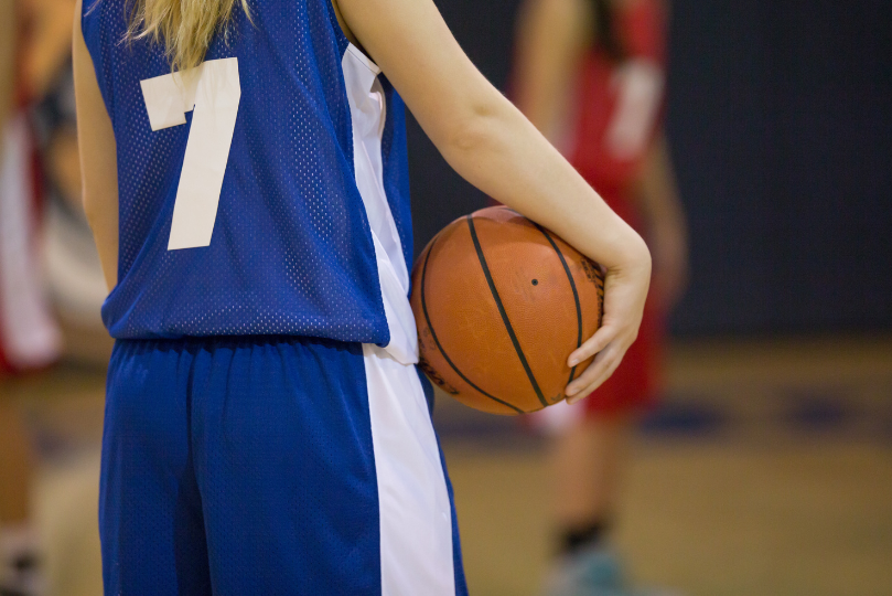 A female basketball player wearing a blue jersey with the number 7 holds a basketball at her side during a game. The background is blurred, showing opposing players in red uniforms on the court.