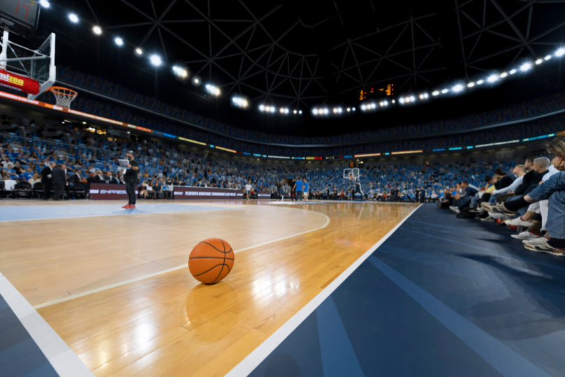 A basketball rests on the polished hardwood court of a large indoor arena, with bright overhead lights illuminating the scene. Players warm up in the background, while a crowd of spectators fills the blue stadium seats.