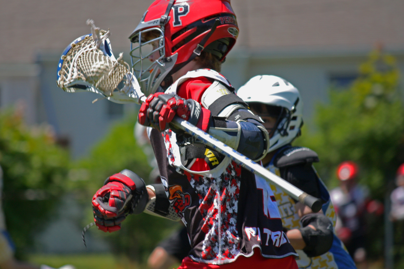 A lacrosse player wearing a red and black helmet with a "P" logo sprints forward, gripping a lacrosse stick with a netted head. He wears protective gear, including gloves and arm pads, while an opposing player in a white helmet closely follows in the background. The scene takes place on a bright, outdoor field.