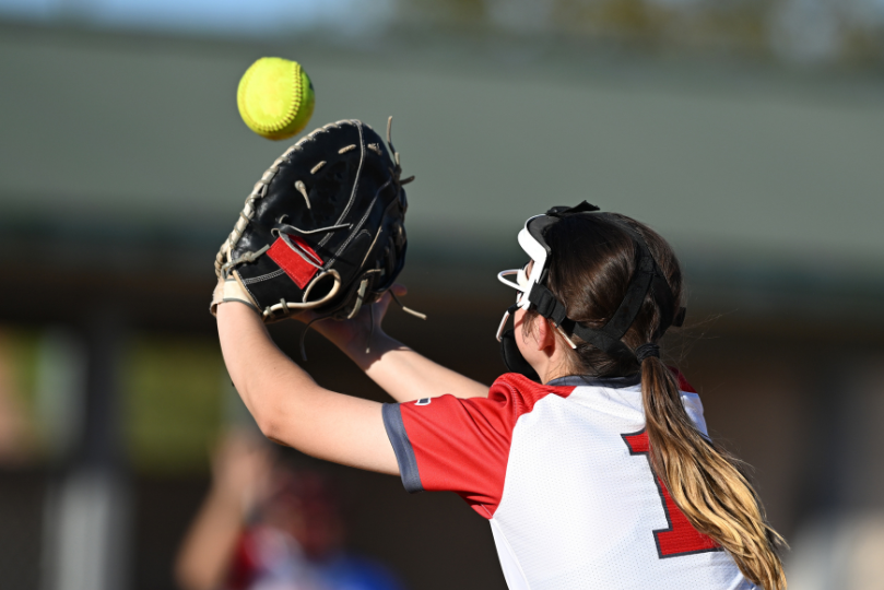 A female softball catcher in a white and red jersey with the number 1 reaches up with her black glove to catch a yellow softball. She wears protective face gear, and the background is slightly blurred, showing a field and spectators.