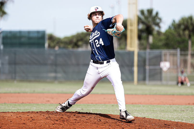 A baseball pitcher in a navy blue Prime jersey with the number 24 stands on the mound, winding up to throw the ball. He wears a white cap with a "P" logo, white pants, and a turquoise baseball glove. The background features a fenced field with trees and a tall yellow foul pole.