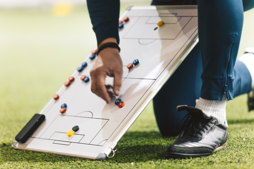 A soccer coach kneels on a grassy field, using a tactical whiteboard with magnetic markers to illustrate game strategies. The coach wears dark athletic pants and black cleats, focusing on positioning and team formation.