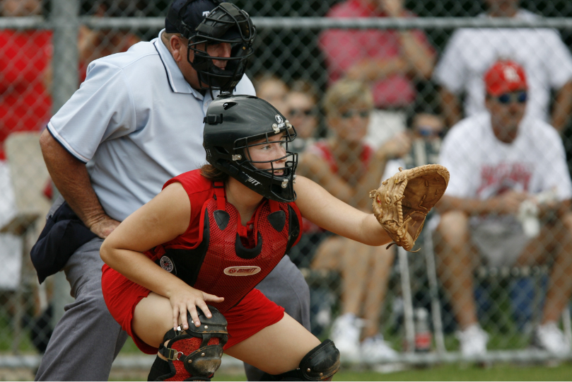 A female softball catcher in red protective gear crouches behind home plate, extending her glove to receive the pitch. An umpire in a light blue shirt and black helmet stands behind her, while spectators watch from the stands behind a chain-link fence.