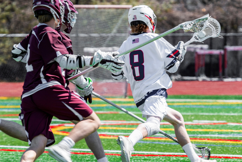 Two lacrosse players in action on a brightly colored artificial turf field. One player in a white jersey with the number 8 is sprinting forward with the ball in his lacrosse stick, while an opposing player in a maroon uniform attempts to defend. Both athletes are wearing helmets, gloves, and protective gear