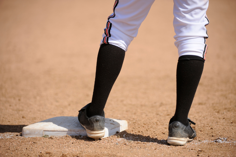 A baseball player wearing white pants with black socks and cleats stands on a base, ready to make a move on the dirt infield. The close-up shot focuses on the player's legs and the base, highlighting the intensity of the game.