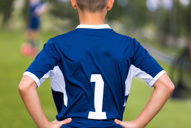 A young soccer player wearing a blue jersey with the number 1 stands with hands on hips, watching the game on a grassy field. The background is blurred, showing another player in action.