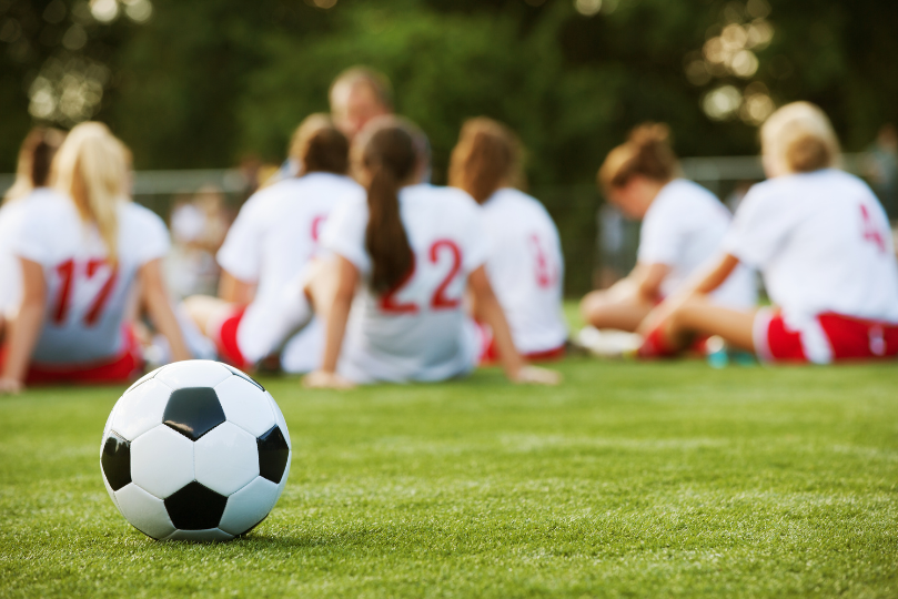 A soccer ball rests on a grassy field in the foreground, while a group of female soccer players in white and red uniforms sit together in the background, listening to their coach during a break.