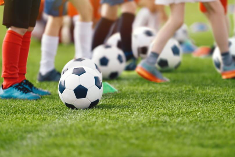 A group of young soccer players stand on a grassy field with soccer balls at their feet, wearing colorful uniforms and cleats. The image focuses on the lower half of their legs, capturing the energy of a youth soccer practice or training session.