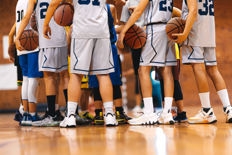 A group of basketball players in white and blue uniforms huddle together on a polished gymnasium floor, holding basketballs. Their legs and sneakers are prominently visible, emphasizing teamwork and preparation before the game.