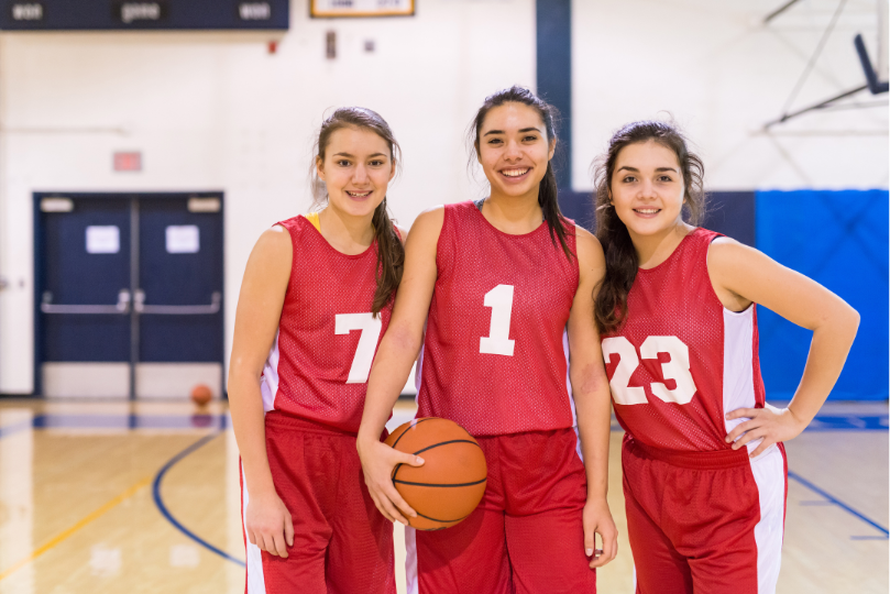 Three female basketball players in red and white uniforms stand together on a gymnasium court, smiling at the camera. The player in the center, wearing the number 1 jersey, holds a basketball, while her teammates, wearing numbers 7 and 23, stand beside her with their arms relaxed.