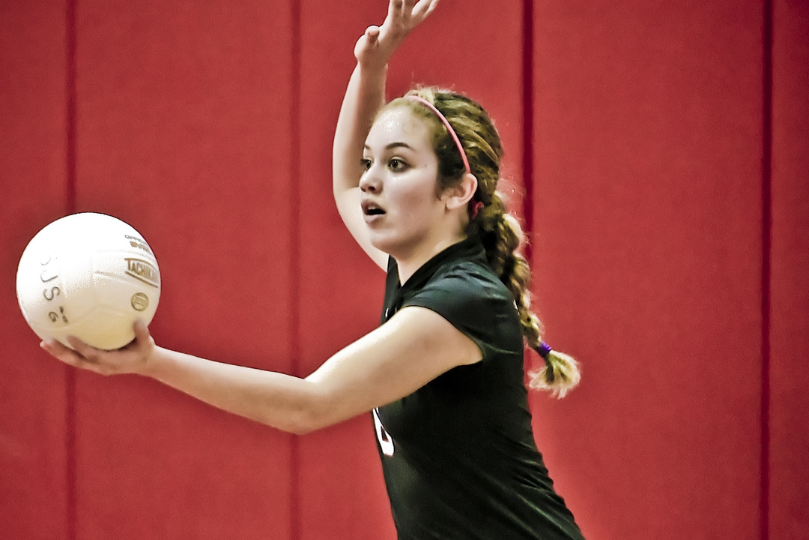 A young female volleyball player in a black jersey prepares to serve, holding a white volleyball in one hand while raising the other. She has a focused expression, braided hair, and a pink headband, with a red gymnasium wall in the background.