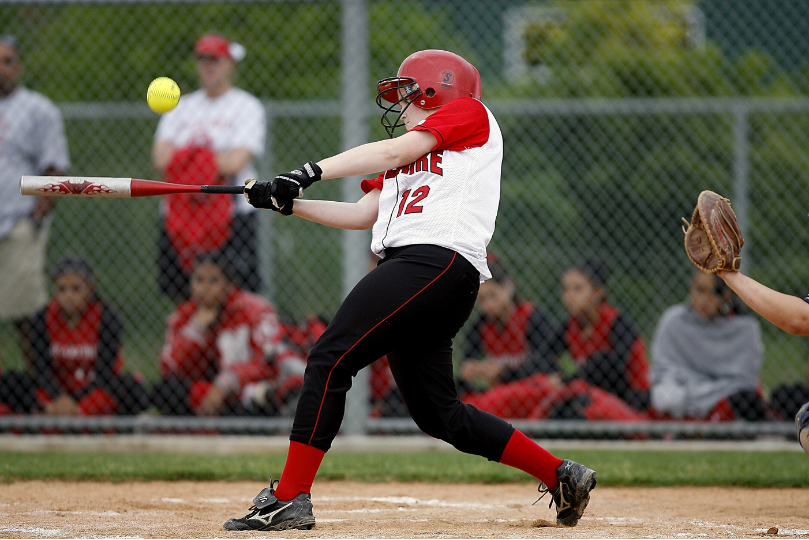 A female softball player in a red helmet and white jersey with the number 12 swings her bat at an incoming yellow softball. She is in a powerful stance at home plate, while a catcher’s glove is visible to the side. Spectators and teammates in red uniforms watch from behind a chain-link fence.